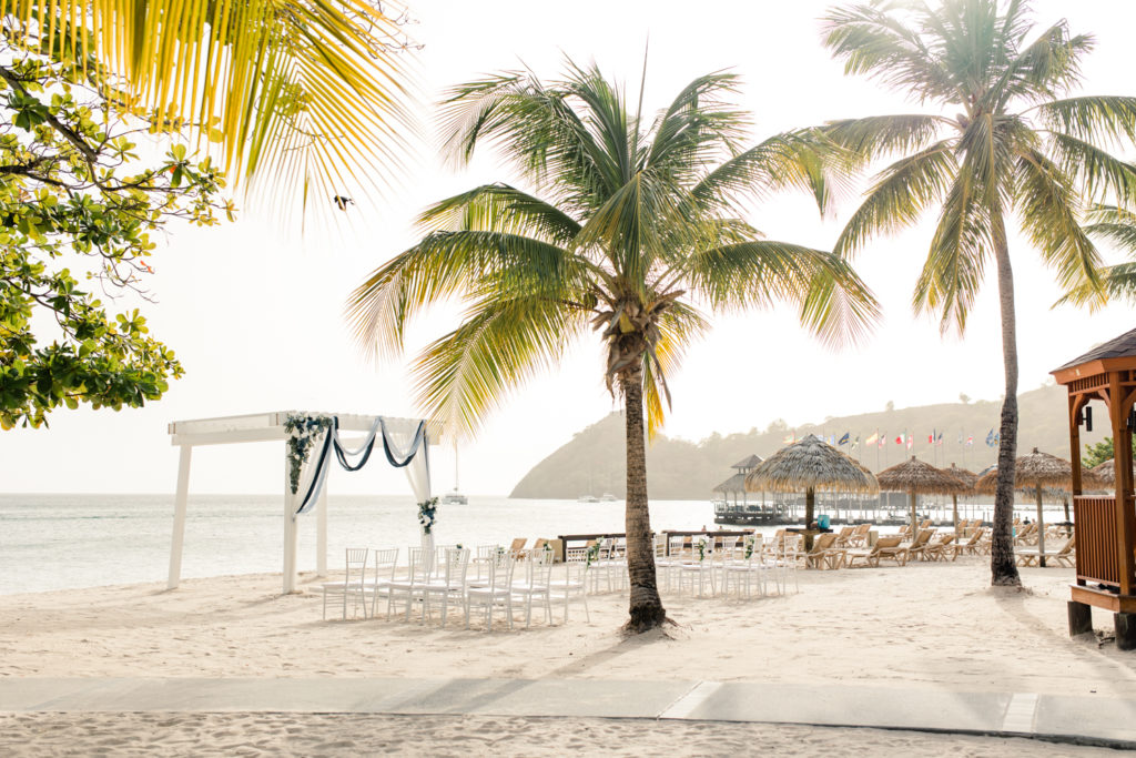 Ceremony set up on the beach of St Lucia with thirty white chairs and arbor surrounded by large palm chairs.