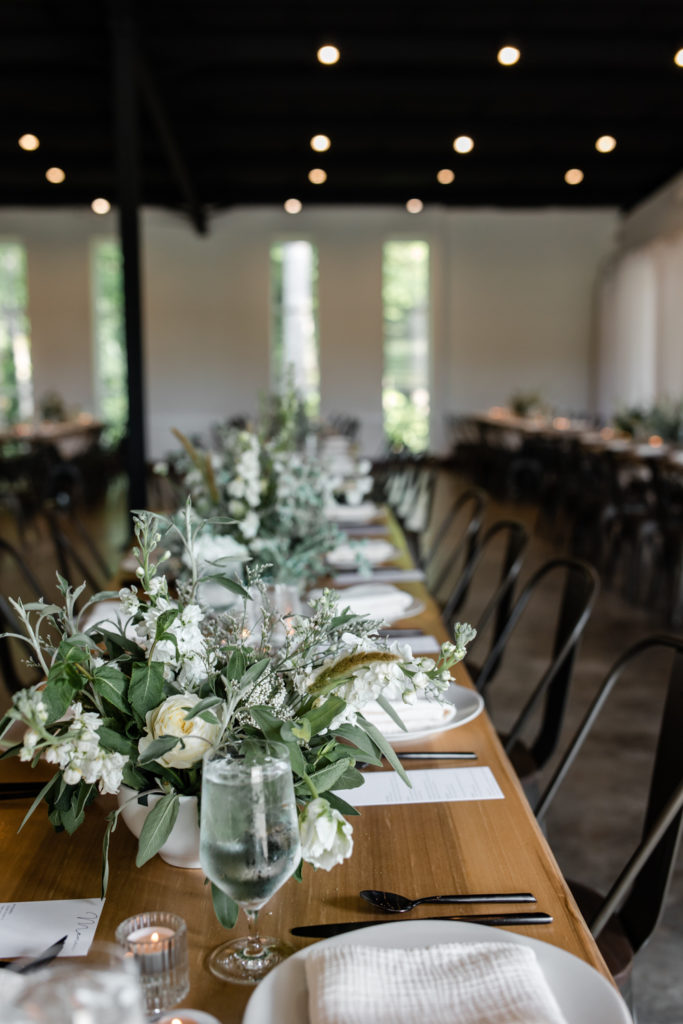 Wedding reception details of wood rustic tables with white and green bouquet table scapes at The Meadows Raleigh wedding venue in Raleigh NC. Photographed by Charlotte wedding photographer.