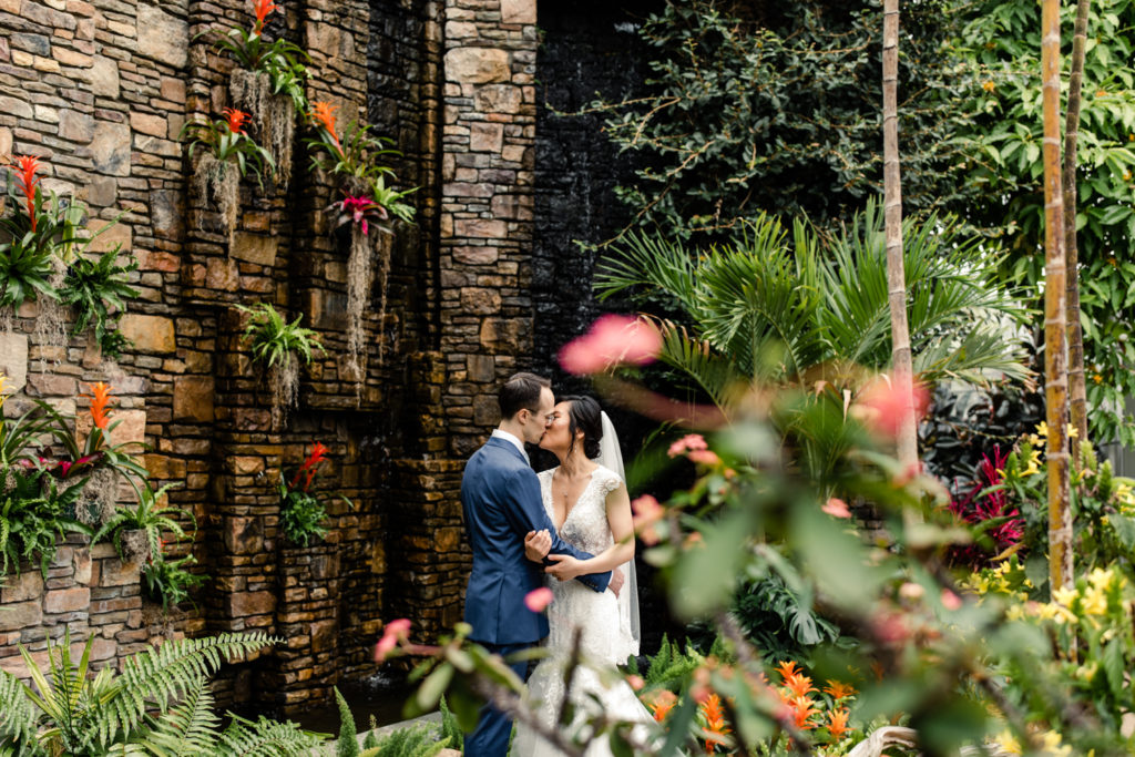 Bride in white lace gown and veil kissing groom with brown hair in blue suit at Daniel Stowe Botanical Gardens in Charlotte photographed by Stephanie Bailey Photography.