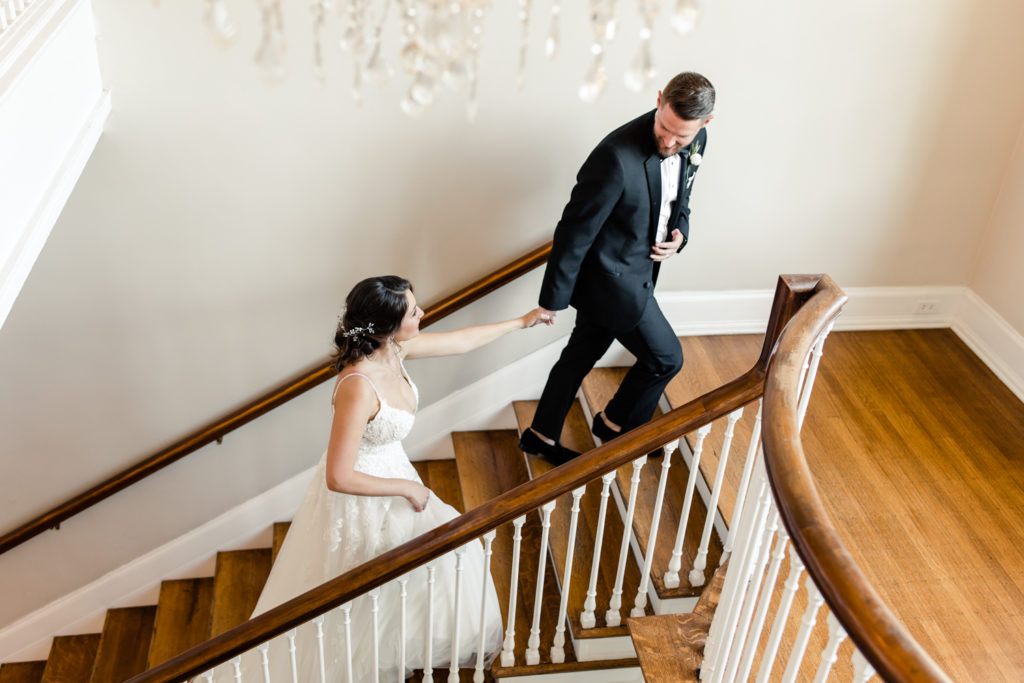 Groom in black suit holding hands with bride in white gown leading her up a brown and white staircase at SePark Mansion Gastonia Wedding Venue. Photographed by Charlotte wedding photographer.