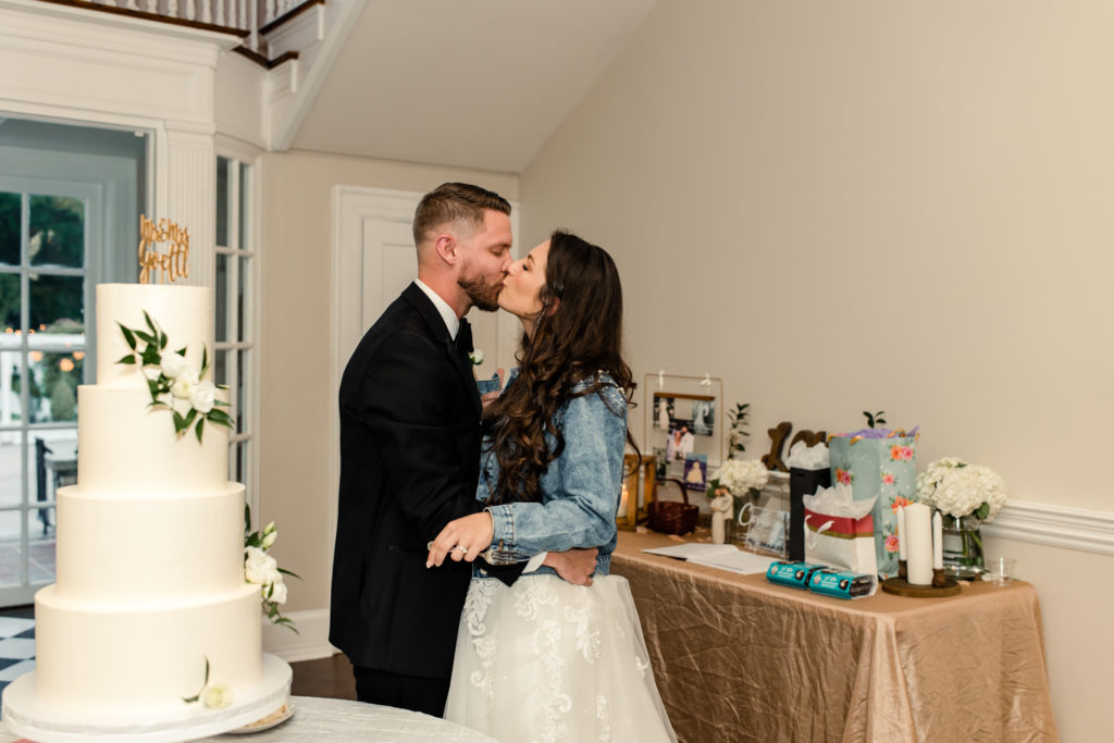 Bride in white dress and denim jacket kissing groom in black suit next to a four layered white cake at SePark Mansion Gastonia Wedding Venue. Photographed by Charlotte wedding photographer.