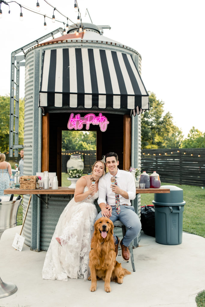Bride in white lace dress sitting with groom in white button down and floral tie holding Ben and Jerry's ice cream cones with golden retriever at Bansen Farms wedding venue in Charlotte NC.