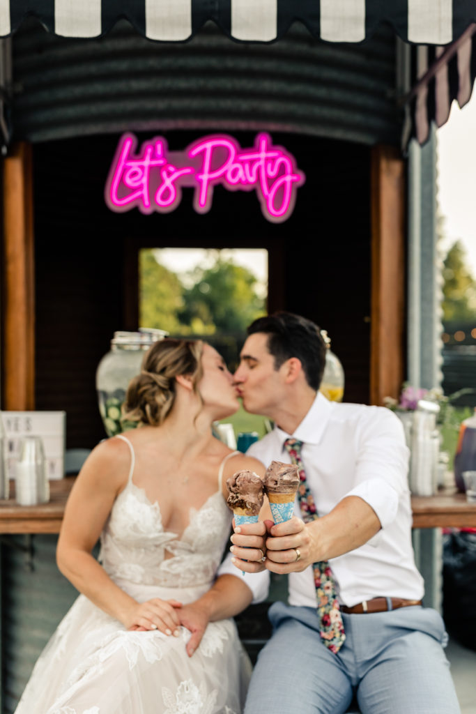 Bride in white lace dress kissing groom in white button down and floral tie holding ice cream cones at Bansen Farms wedding venue in Charlotte NC.