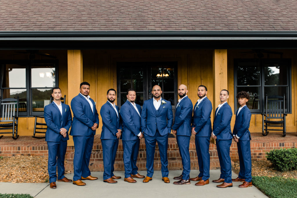 Groom standing with hands in pockets with seven groomsmen in blue suits at The Meadows Raleigh wedding venue in Raleigh NC. Photographed by Charlotte wedding photographer.