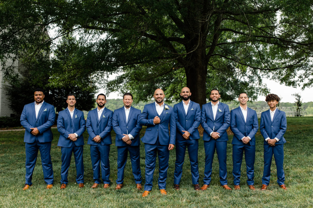 Groom standing with seven groomsmen in blue suits at The Meadows Raleigh wedding venue in Raleigh NC. Photographed by Charlotte wedding photographer.