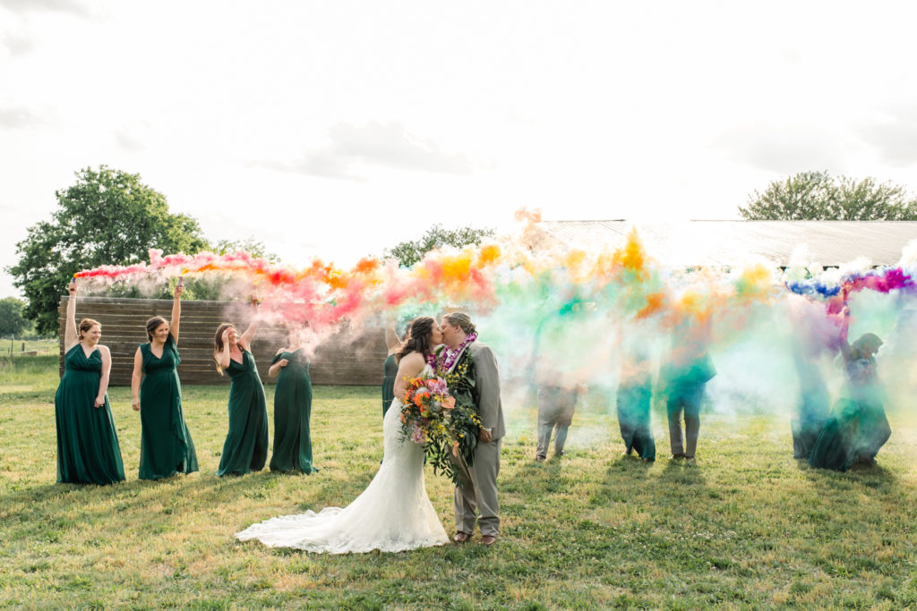 Bride, in white lace dress, kissing her bride, in tan suit, under rainbow smoke bombs at 1932 Barn Wedding Venue in Charlotte