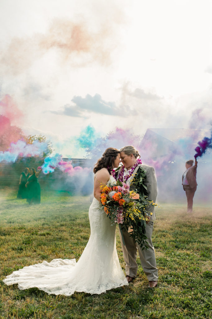 Bride, in white lace dress, nuzzling with her bride, in tan suit, under rainbow smoke bombs at 1932 Barn Wedding Venue in Charlotte