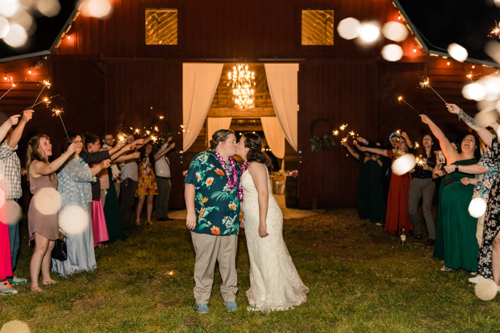 Bride, in green Hawaiian shirt kissing her bride in white lace dress during wedding sparklier exit in front of the red barn Wedding reception tables at 1932 Barn Wedding Venue in Charlotte