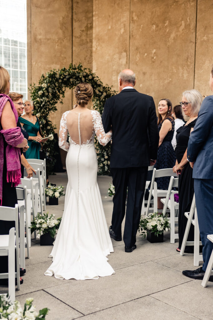 Bride, in white dress, holding white bouquet, walking down aisle with father, in black suit, at the Mint Museum wedding venue in Uptown Charlotte