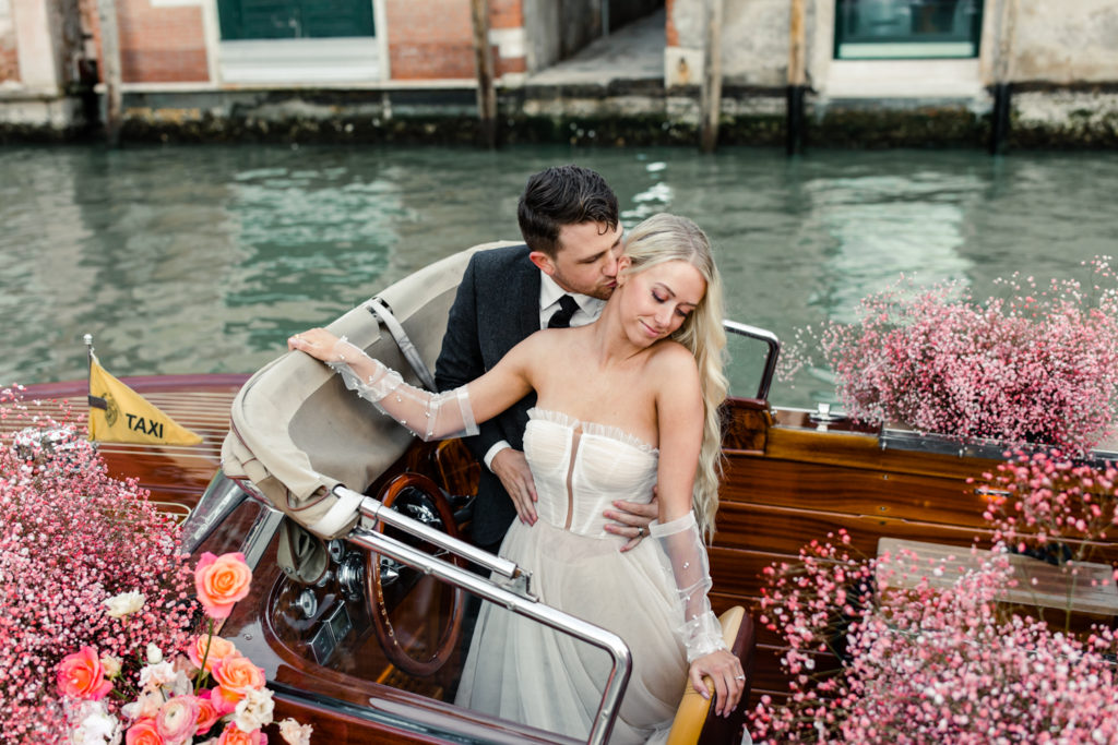 Groom kissing the cheek of bride in water taxi decorated in pink flowers in Venice Italy. Photographed by Charlotte wedding photographer.