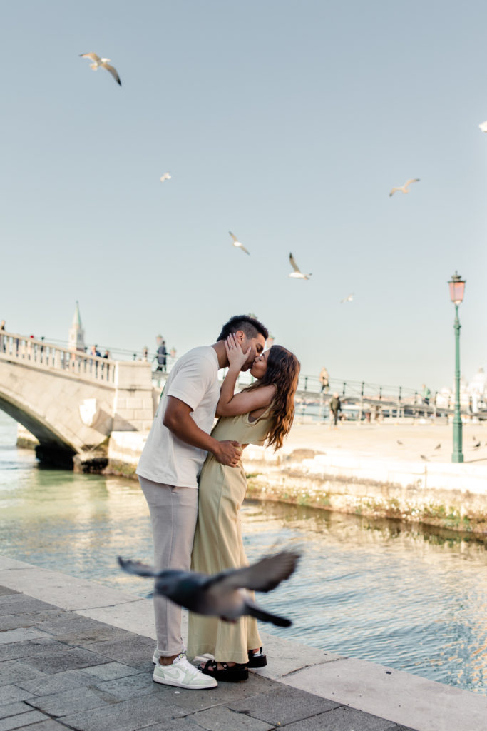 Boy, in white shirt, kissing girl, in yellow jumpsuit, as pigeons fly around them in Venice Italy. Photographed by Charlotte wedding photographer.