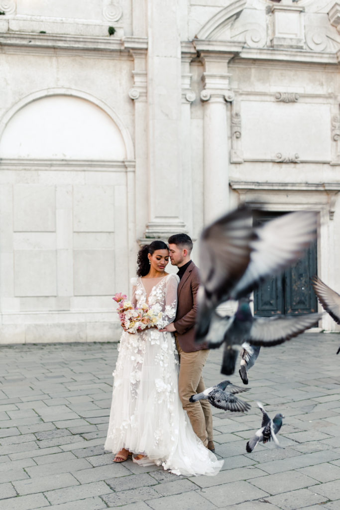 Groom, in brown suit, standing with bride, in white gown and pink bouquet, in Venice Italy while pigeon fly. Photographed by Charlotte wedding photographer. 