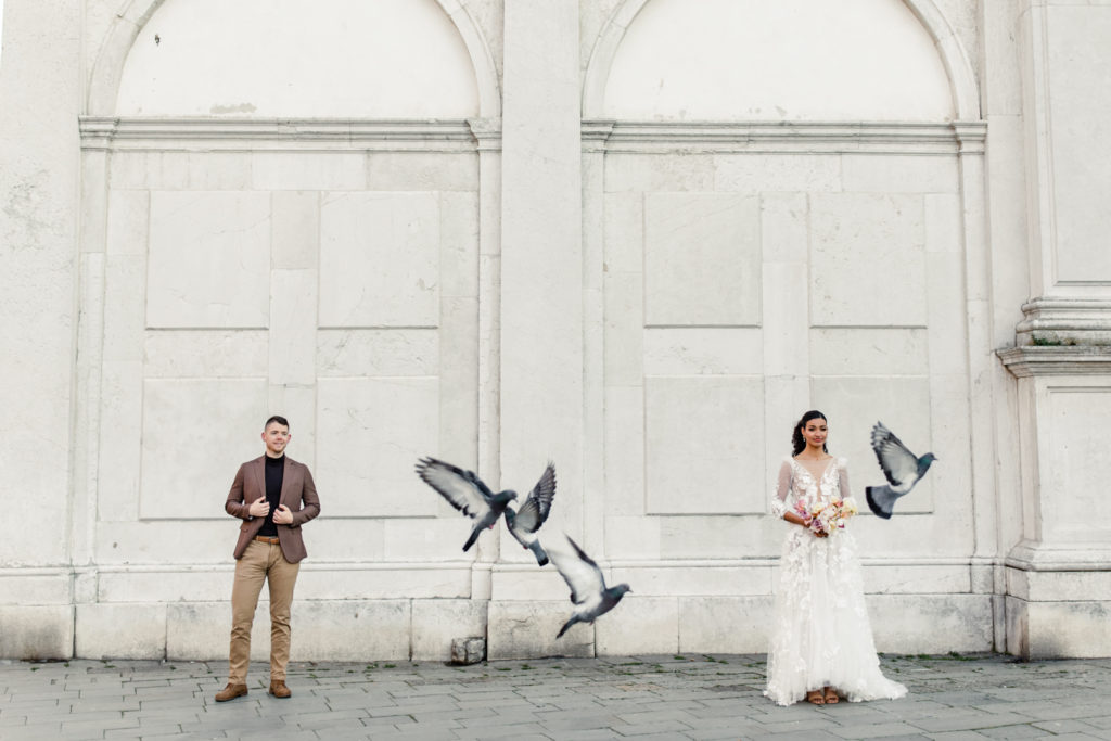 Groom, in brown suit, standing with bride, in white gown and pink bouquet, in Venice Italy while pigeon fly. Photographed by Charlotte wedding photographer. 