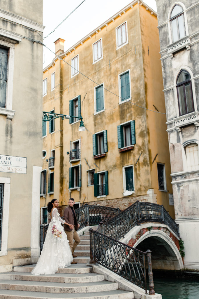 Groom, in brown suit, walking up bridge with bride, in white gown, in front of yellow building in Venice Italy. Photographed by Charlotte wedding photographer. 