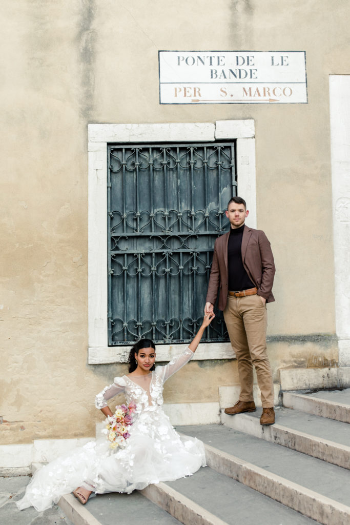 Groom, in brown suit, holding hands with bride, in white gown, on stairs of bridge in front of yellow building in Venice Italy. Photographed by Charlotte wedding photographer. 