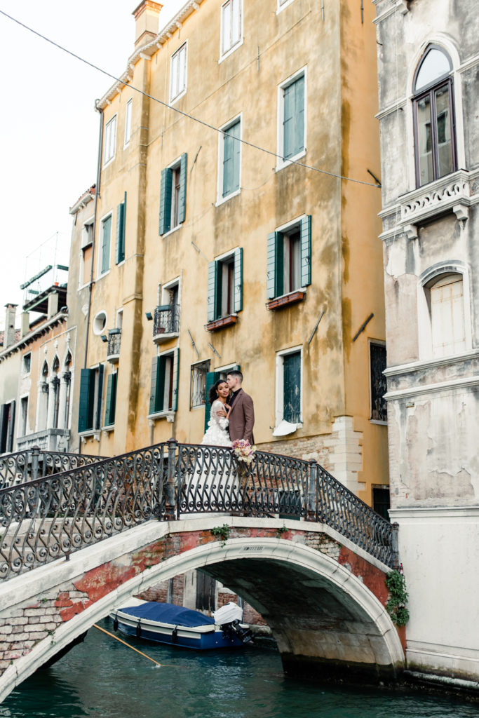 Groom, in brown suit, kissing the cheek of bride, in white gown, on a bridge in front of yellow building in Venice Italy. Photographed by Charlotte wedding photographer. 