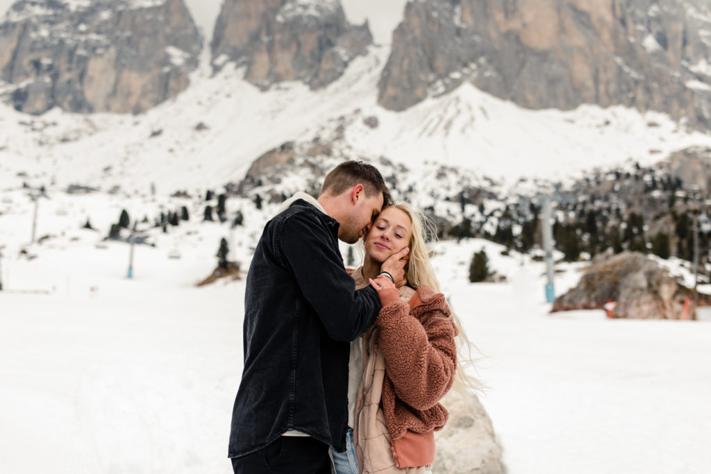 Boy, in black jacket, kissing girl on the cheek in the snow in Dolomites Italy. Photographed by Charlotte wedding photographer.