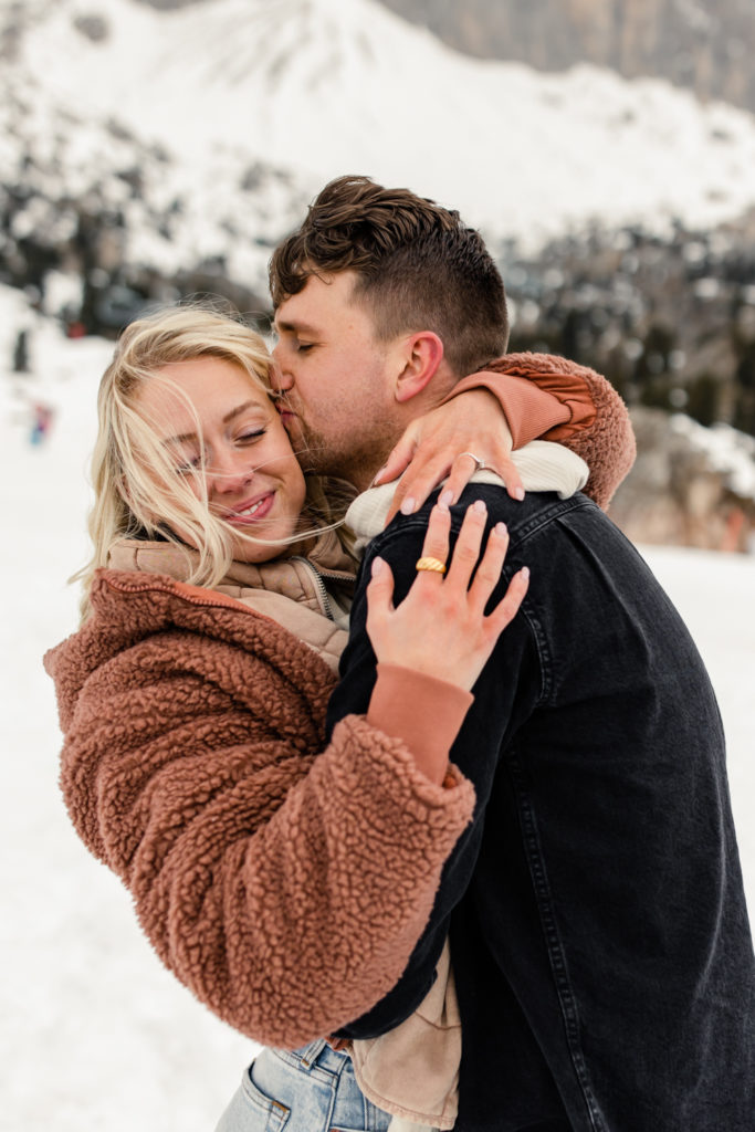 Boy, in black jacket, hugging and kissing girl on the cheek in the snow in Dolomites Italy. Photographed by Charlotte wedding photographer.