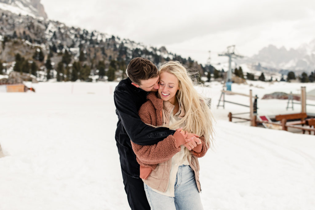 Boy, in black jacket, hugging and kissing girl on the cheek in the snow in Dolomites Italy. Photographed by Charlotte wedding photographer.