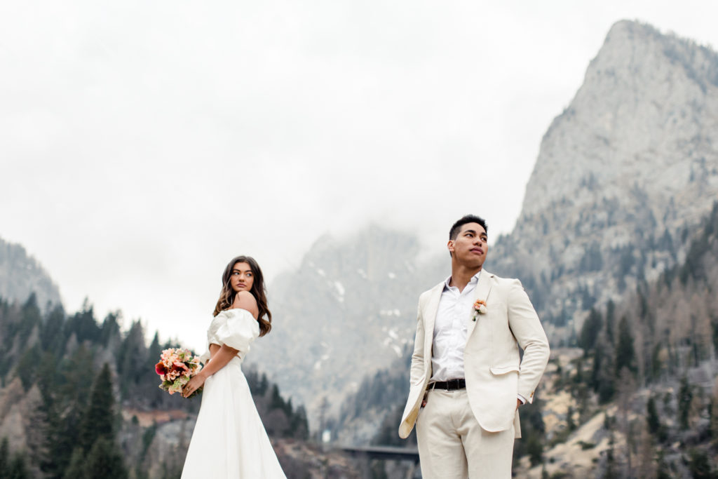 Groom, in cream suit, standing with bride, in white dress holding pink bouquet, on a mountain in Dolomites Italy. Photographed by Charlotte Wedding Photographer.