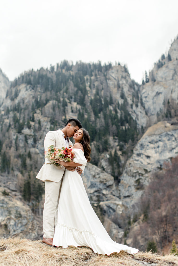 Groom, in cream suit, kissing bride, in white dress holding pink bouquet in Dolomites Italy. Photographed by Charlotte Wedding Photographer.