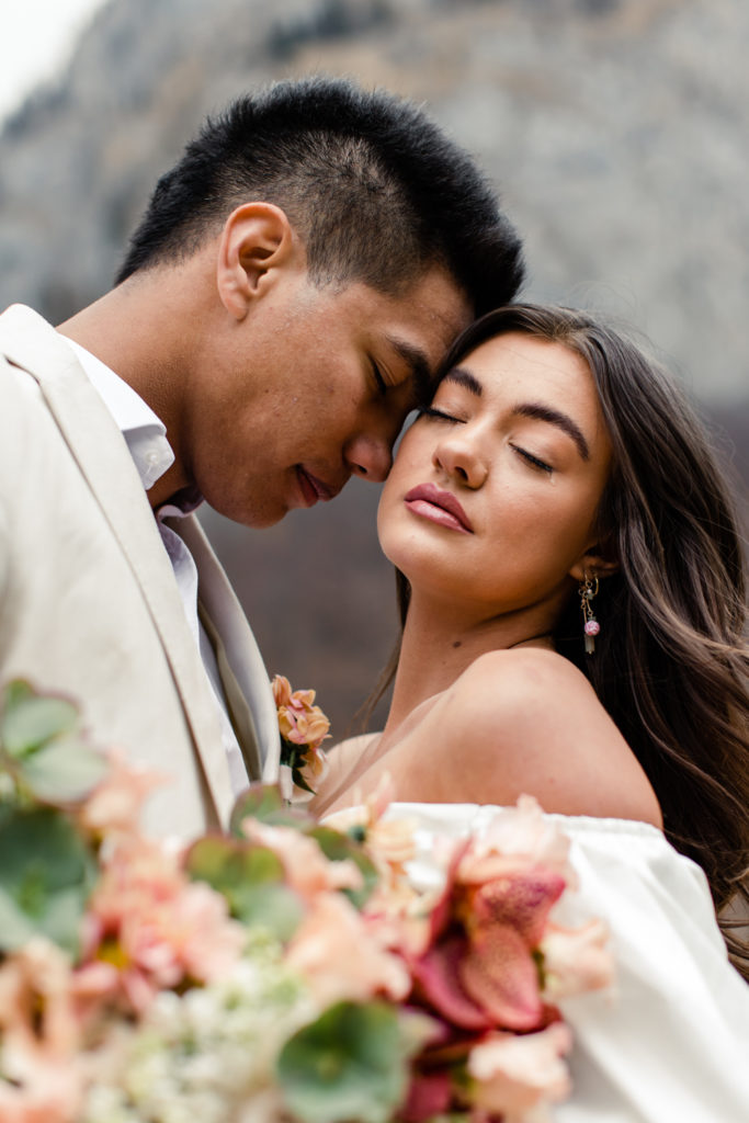 Groom, in cream suit, nuzzling the bride, in white dress holding pink bouquet in Dolomites Italy. Photographed by Charlotte Wedding Photographer.