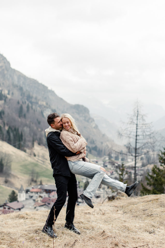 Boy, in black jacket and pants, picking up girl and spinning her around in Dolomites Italy. Photographed by Charlotte wedding photographer.