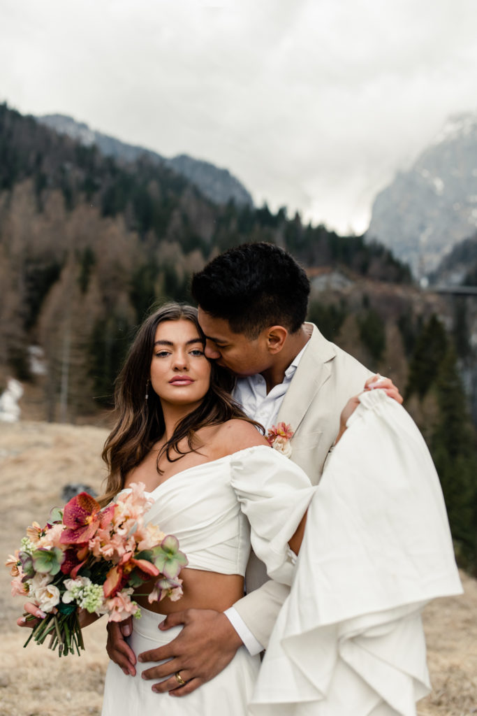Groom, in cream suit, kissing bride, in white dress holding pink bouquet in Dolomites Italy. Photographed by Charlotte Wedding Photographer.