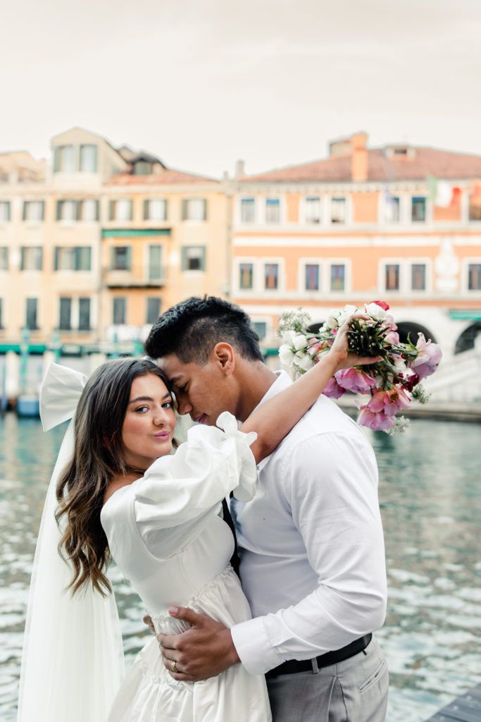 Bride, in white gown and veil, hugging groom holding pink bouquet in Venice Italy. Photographed by Charlotte wedding photographer. 