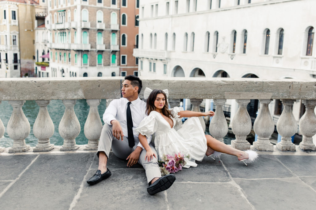 Bride, in white gown and brown sunglasses, sitting with groom in Venice Italy. Photographed by Charlotte wedding photographer. 