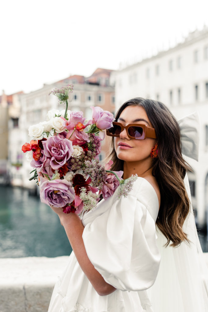 Bride, in white gown and veil with brown sunglasses, holding pink and purple bouquet in Venice Italy. Photographed by Charlotte wedding photographer. 