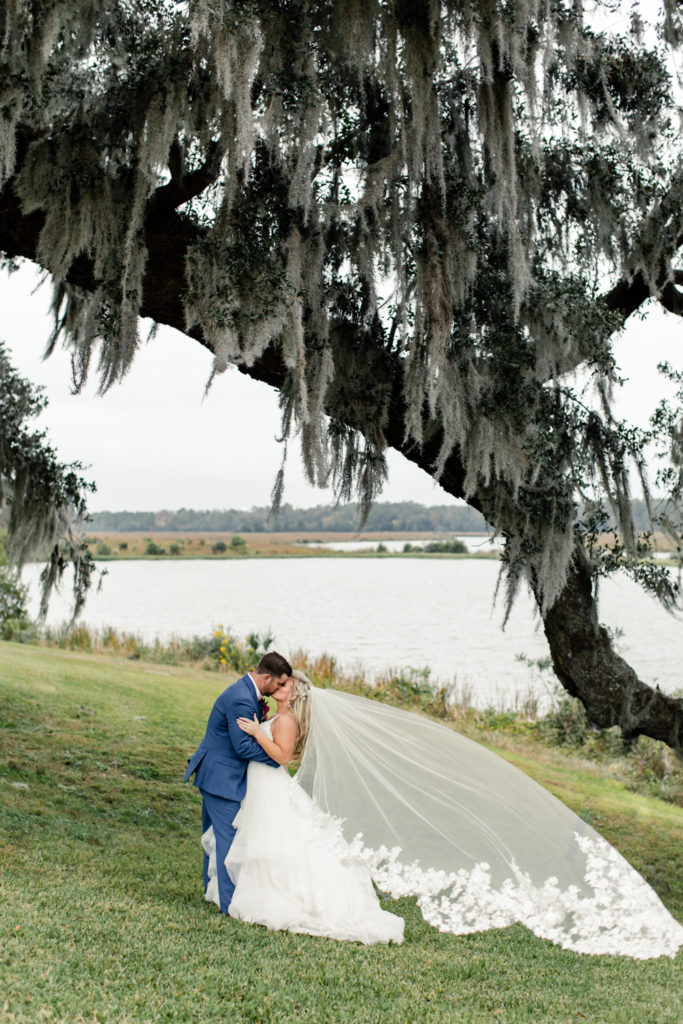 Bride, in white dress with long lace veil, kissing groom, in blue suit, under a large oak tree at Middleton Place wedding venue in Charleston. Photographed by Charlotte Wedding Photographer.