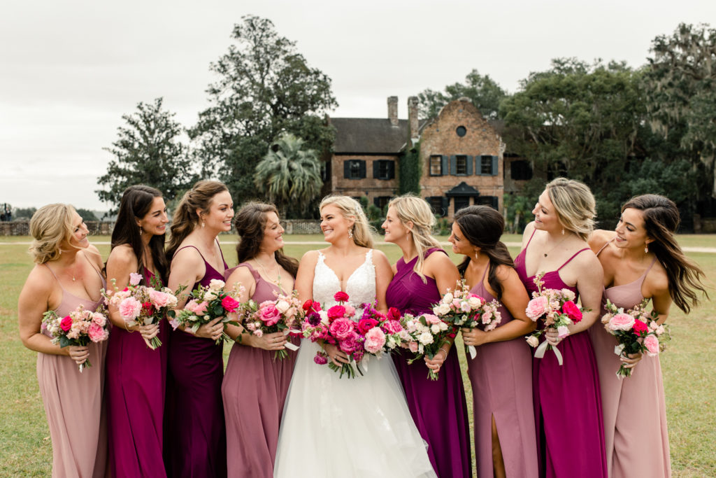 Bride, in white dress with blonde hair, laughing with her eight bridesmaids in pink dresses, all holding pink bouquets at Middleton Place wedding venue in Charleston. Photographed by Charlotte Wedding Photographer.