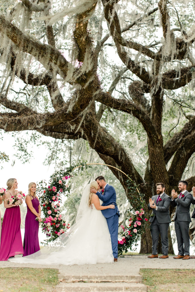 Bride, in white wedding gown and veil, kissing groom during ceremony, in blue suit, in front of a gold arbor with pink florals under a giant oak tree at Middleton Place wedding venue in Charleston. Photographed by Charlotte Wedding Photographer.