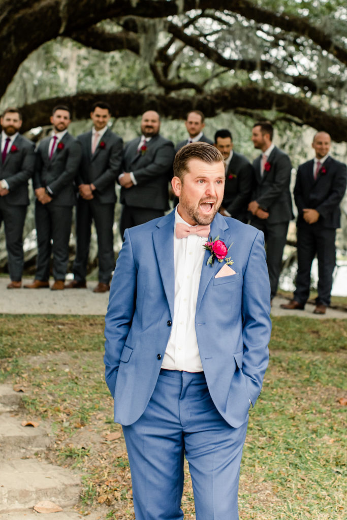 Groom, with brown hair and blue suit and pink bow tie with pink boutonnière, sees bride for the first time walking down aisle at Middleton Place wedding venue in Charleston. Photographed by Charlotte Wedding Photographer.