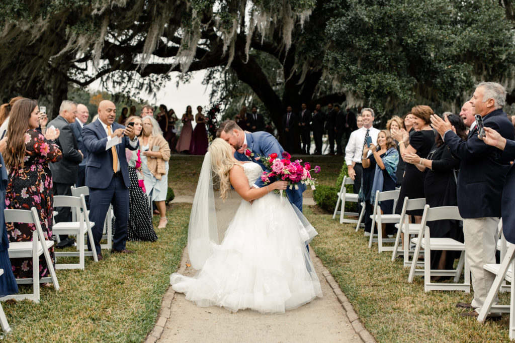 Bride, in white wedding gown and veil, kissing groom during ceremony, in blue suit, under a giant oak tree at Middleton Place wedding venue in Charleston. Photographed by Charlotte Wedding Photographer.