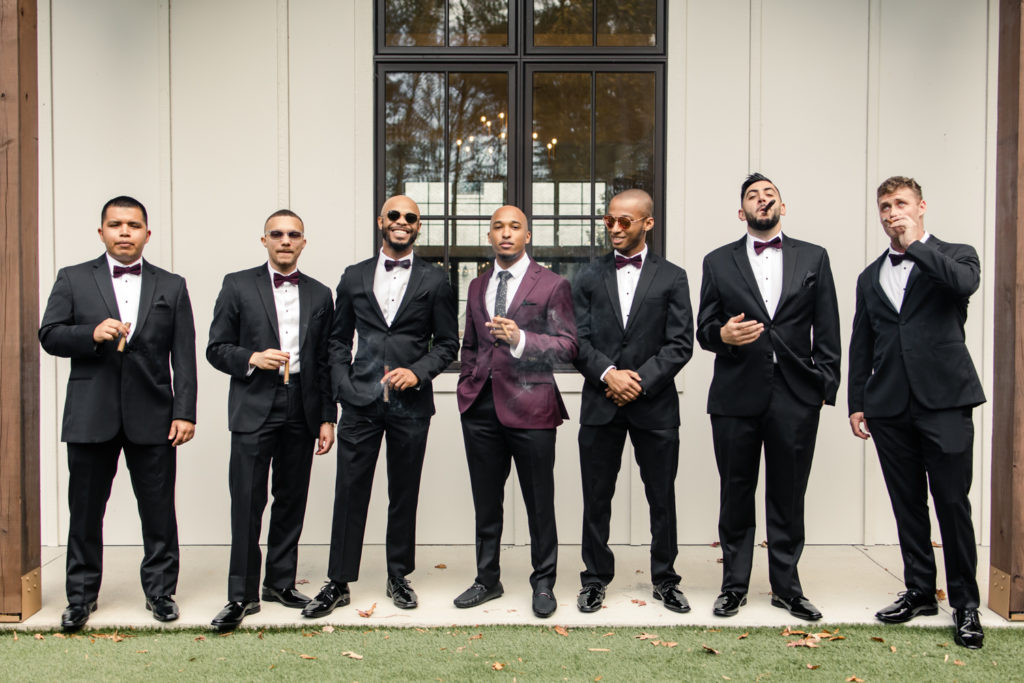 Groom, in burgundy suit, smoking cigars with six groomsmen at the Bradford wedding venue in Raleigh NC.