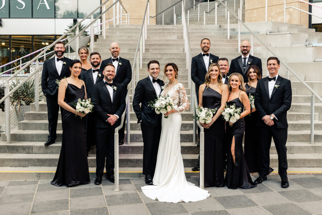 Bride, in white lace dress, standing with groom, in black suit, on a staircase with the wedding party, dressed in black, at the Mint Museum wedding venue in Uptown Charlotte