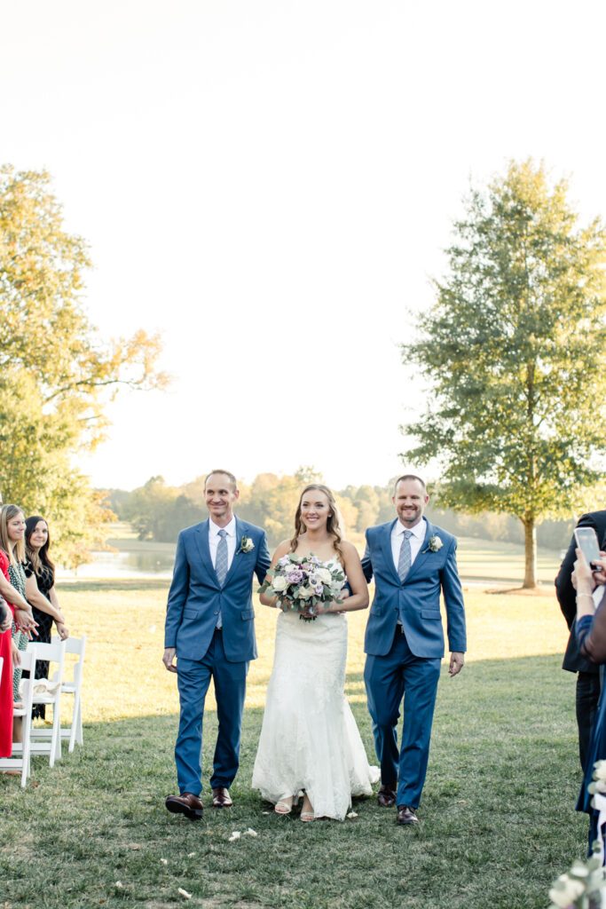 Bride, in white lace dress, walking down aisle with two brothers, dressed in blue suits, at Long View Country Club wedding venue in Charlotte NC