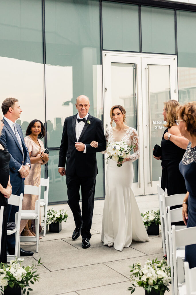 Bride, in white dress, holding white bouquet, walking down aisle with father, in black suit, at the Mint Museum wedding venue in Uptown Charlotte