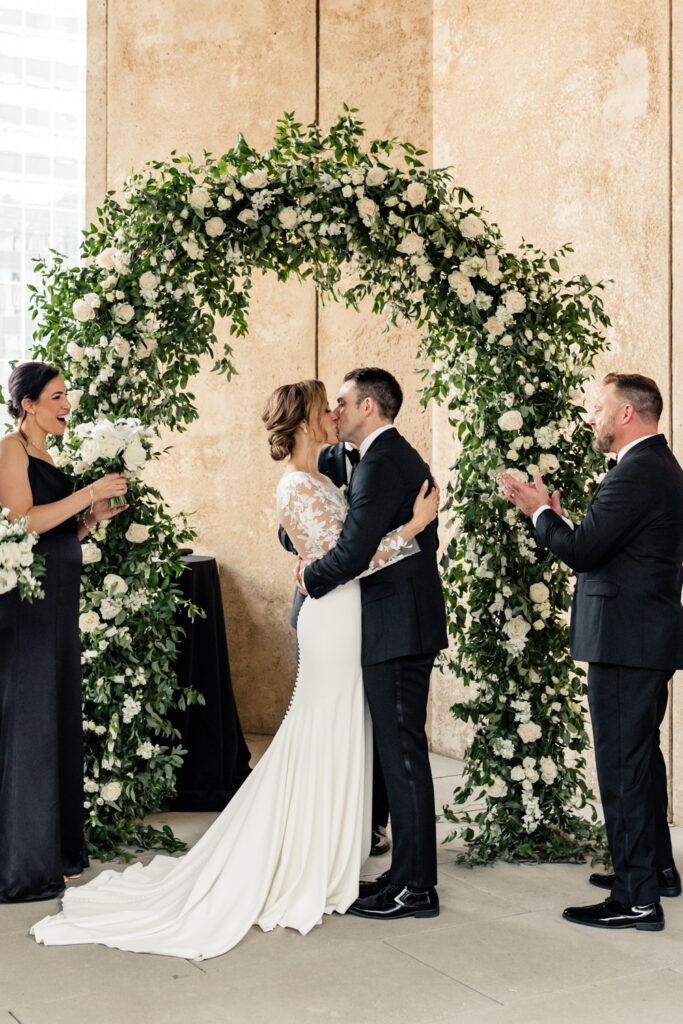 Bride, in white dress, kissing the groom, in black suit, for the first time during ceremony, under a white and green floral arch, at the Mint Museum wedding venue in Uptown Charlotte