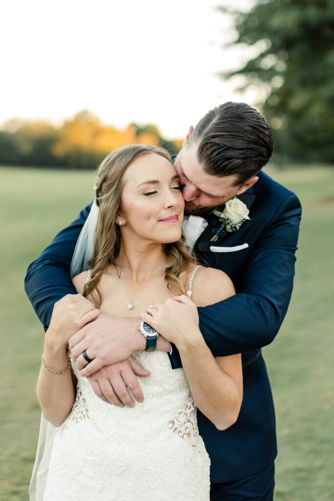 Groom, in navy suit, kissing the cheek of bride, in white dress, on the golf course at Long View Country Club wedding venue in Charlotte NC