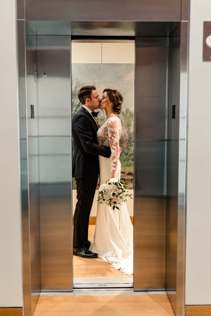 Bride, in white lace dress, kissing groom, in black suit, in the elevator as the doors close at the Mint Museum wedding venue in Uptown Charlotte