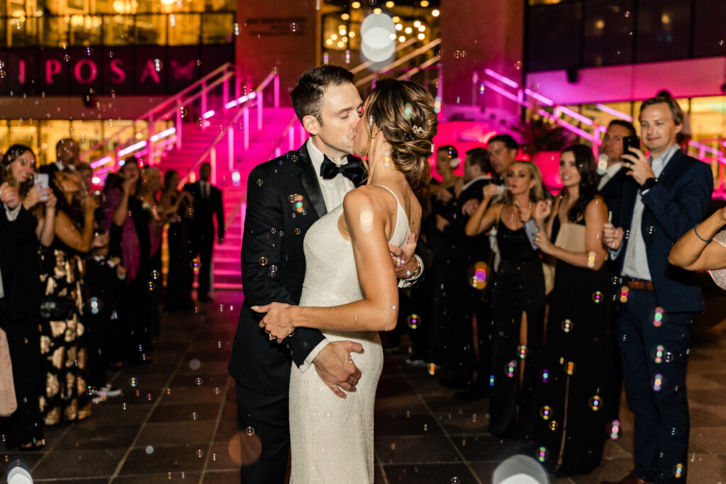 Bride, in a white one shoulder dress, kissing groom, in black suit and bowtie, during reception  bubble exit with at the Mint Museum in Uptown Charlotte.