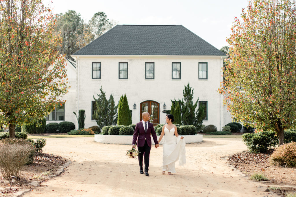 Bride, in white lace gown, walking with groom, in burgundy suit, in front of the Bradford wedding venue in Raleigh NC.