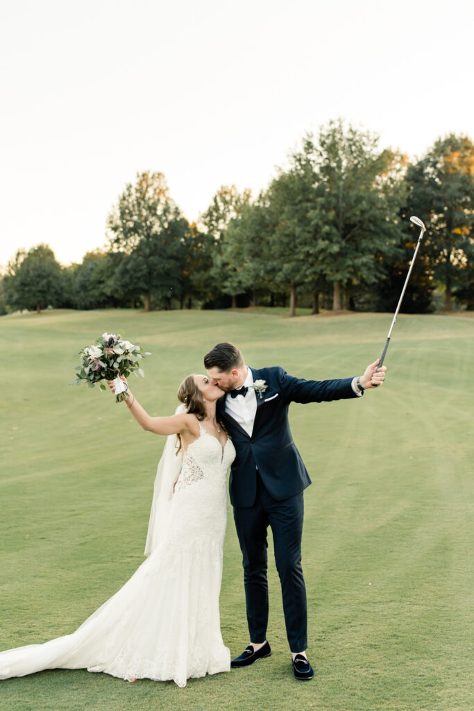 Groom, in navy suit, kissing bride, in white dress, on the golf course while putting her bouquet and his golf club in the air at Long View Country Club wedding venue in Charlotte NC