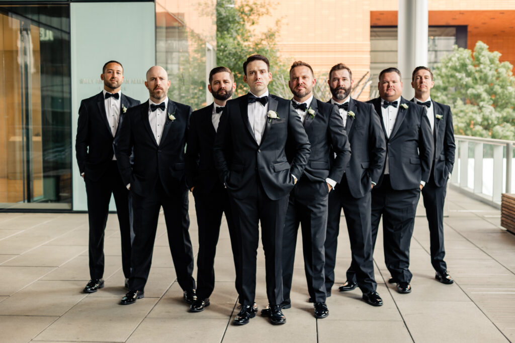 Groom standing with seven groomsmen all dressed in black suits and bowties at the Mint Museum wedding venue in Uptown Charlotte