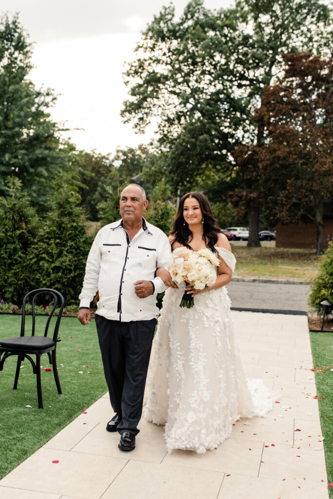 Bride, in white dress and holding pink and white flowers, walks down ceremony aisle with dad at The Highlawn Wedding venue in New Jersey. Photographed by Charlotte Wedding photographer.