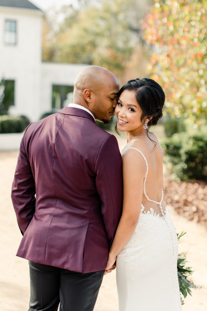 Groom, in burgundy suit, nuzzling the cheek of his bride, in white lace dress, at the Bradford wedding venue in Raleigh NC.