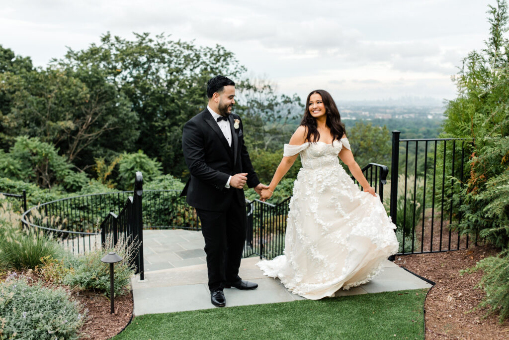 Bride, in white lace dress, holding hands with groom, in black suit, smiling at The Highlawn Wedding venue in New Jersey. Photographed by Charlotte Wedding photographer.